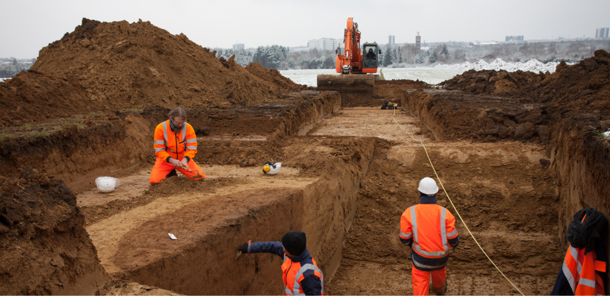 © Seine et Yvelines Archéologie / EPI Yvelines-Hauts-de-Seine Travailleurs en vêtements de sécurité orange sur un chantier de fouilles archéologiques, avec une pelleteuse en arrière-plan et des tranchées creusées dans le sol.